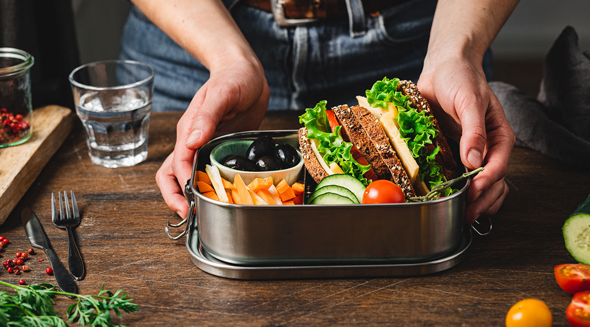 Jemand bereitet eine Lunchbox mit belegten Vollkornbroten, Gemüse und Obst zu 