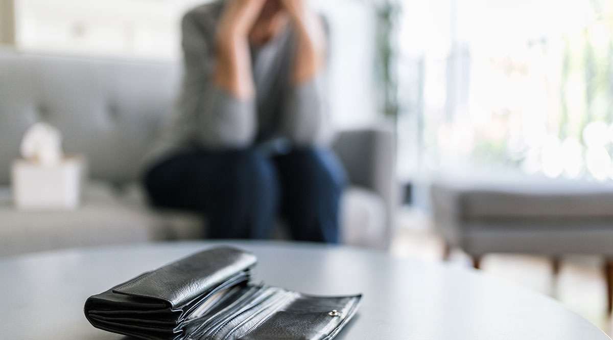 Jemand sitzt verzweifelt auf einem Sofa, im Vordergrund eine geöffnete Geldbörse auf einem dem Tisch