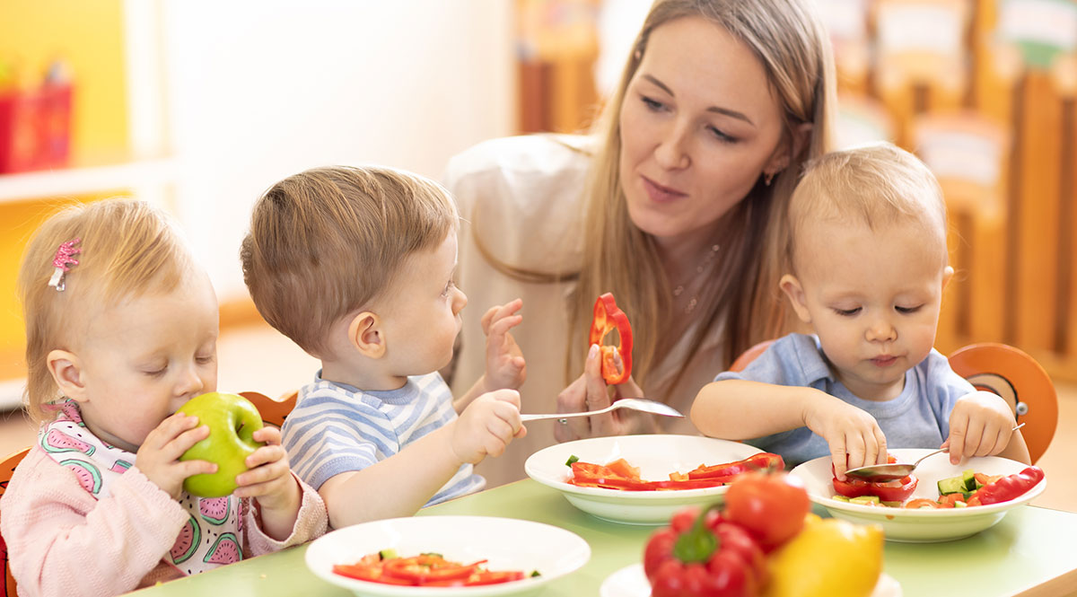 Eine Betreuerin sitzt mit Kleinkindern am Esstisch