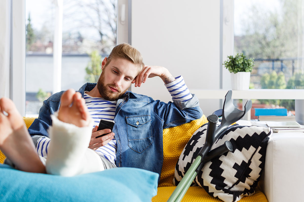 Junger Mann mit gebrochenem Bein auf dem Sofa, Smartphone in der Hand