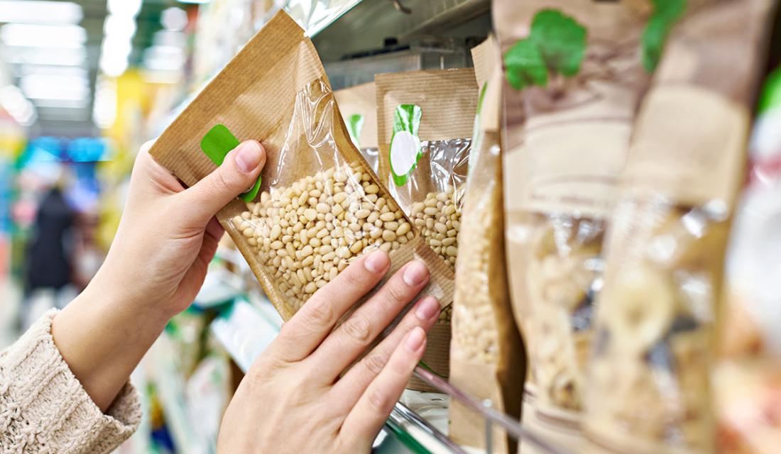 Hände greifen im Regal nach einer Tüte Pinienkerne, in Papierverpackung mit einem grünen Label