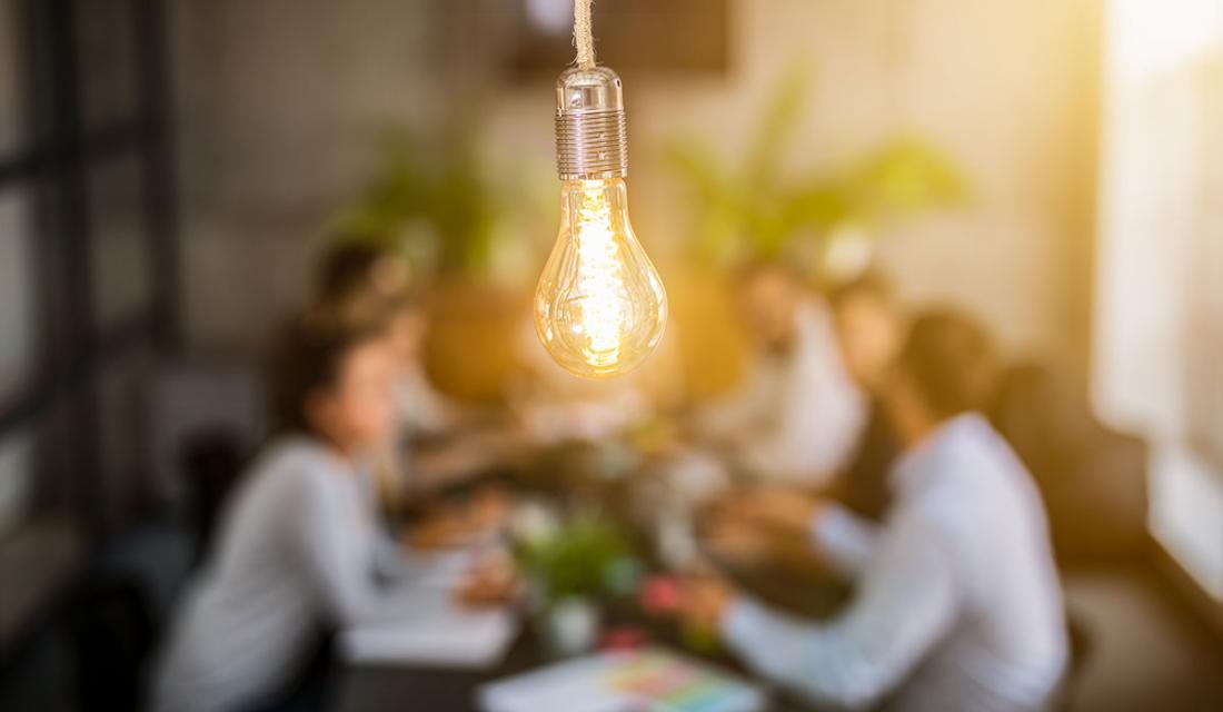 Eine Gruppe Menschen sitzt um einen Tisch und diskutiert, im Vordergrund symbolisiert eine Glühbirne die Ideen