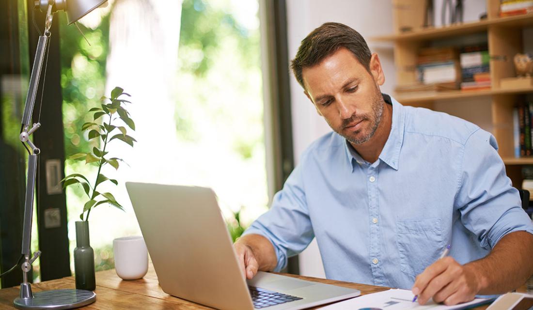 Ein Mann sitzt in einem hellen Arbeitszimmer am Schreibtisch vor einem Laptop und macht Notizn