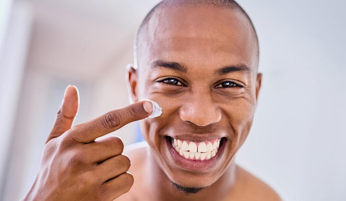 Ein lachender junger Mann trägt Gesichtscreme auf sein Gesicht auf. 