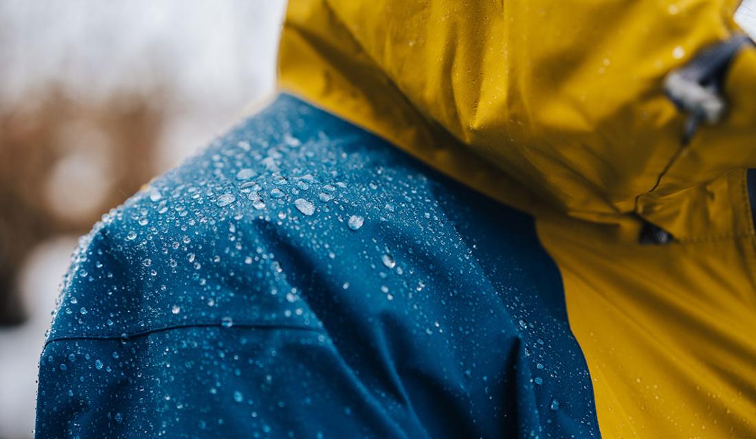Jemand trägt eine blau-gelbe wasserabweisende Regenjacke mit abperlenden Regentropfen auf der Schulter