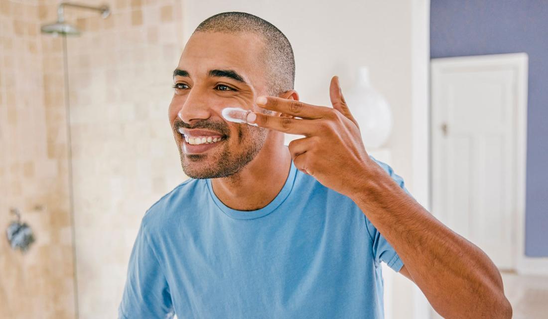 Ein junger Mann steht vor dem Badezimmerspiegel und trägt Gesichtscreme auf seine Wange auf