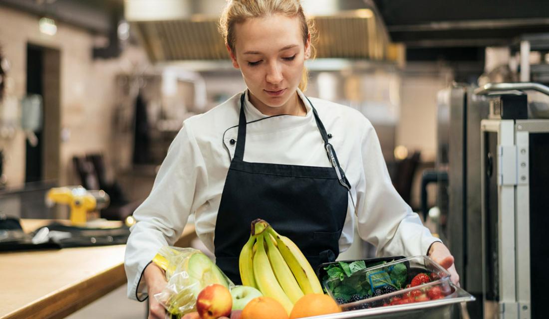Eine Köchin in einer Großküche trägt ein Tablett mit Obst