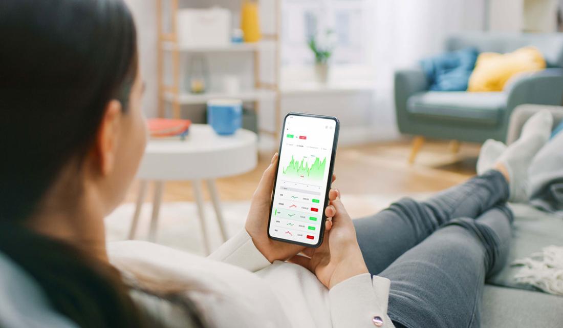 Eine Frau sitzt auf dem Sofa und betrachtet auf dem Smartphone Aktienkurse