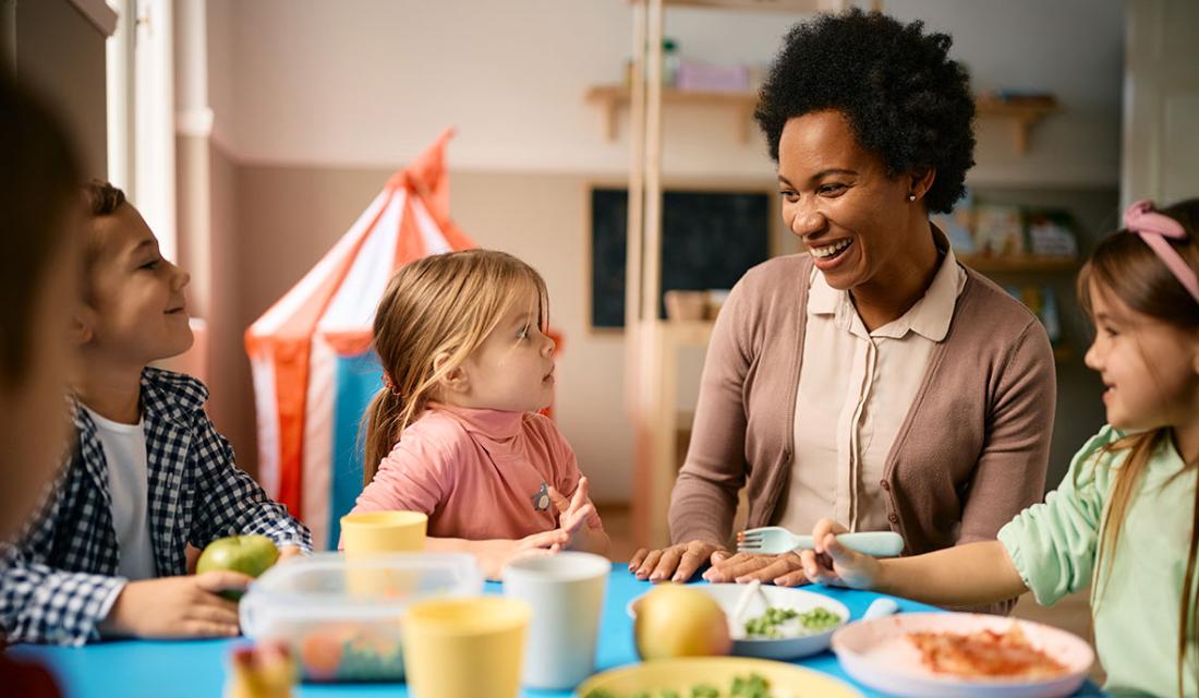Eine Erzieherin sitzt mit Kindern im Kindergartenalter am Esstisch 