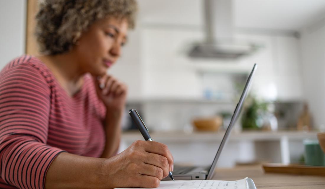 Eine Frau mittleren Alters sitzt in einer Küche am Laptop und macht Notizen auf einem Zettel