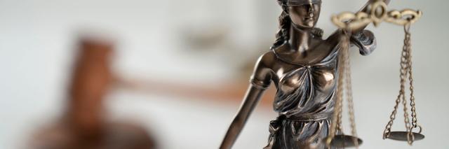 Eine Skulptur der Justitia auf einem Tisch, im Hintergrund der Hammer des Gerichts