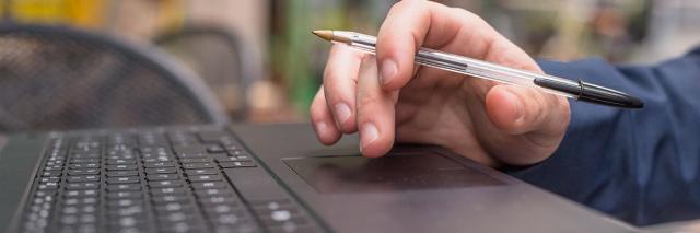 Eine Hand hält einen Kugelschreiber, darunter eine Laptop-Tastatur