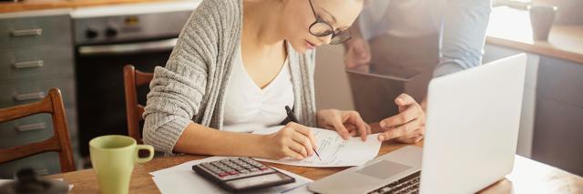Eine Frau sitzt mit Laptop, Taschenrechner, Rechnungen und Stift am Küchentisch, ein Mann steht neben ihr und beugt sich auf eine Rechnung blickend zu ihr herunter.
