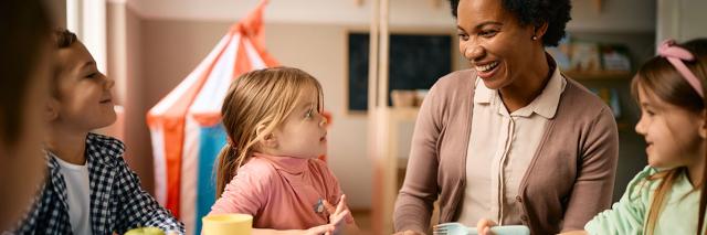 Eine Erzieherin sitzt mit Kindern im Kindergartenalter am Esstisch 