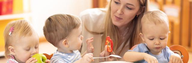 Eine Betreuerin sitzt mit drei Kleinkindern am Esstisch, auf dem Teller mit Gemüse stehen