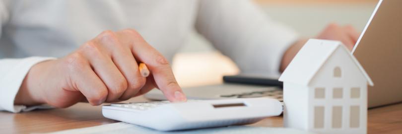 Jemand sitzt an seinem Laptop und rechnet mit einem Taschenrechner. Daneben steht ein kleines Modell eines Hauses.