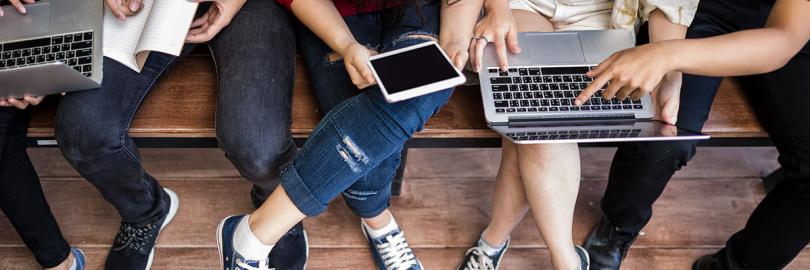 Junge Menschen sitzen auf einer Bank, Laptops und Tablets auf dem Schoß. Man sieht nur ihre Beine. 