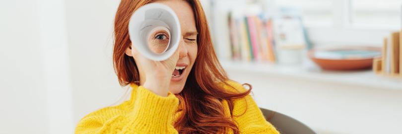 Eine junge Frau im gelben Pullover schaut durch ein zum Fernrohr gerolltes Papier