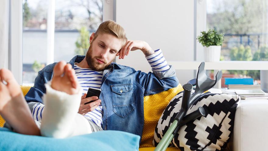 Junger Mann mit gebrochenem Bein auf dem Sofa hält ein Smartphone in der Hand