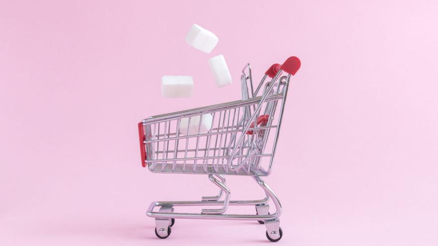 Miniatur-Einkaufswagen vor rosa Hintergrund, ZUckerwürfel fallen hinein