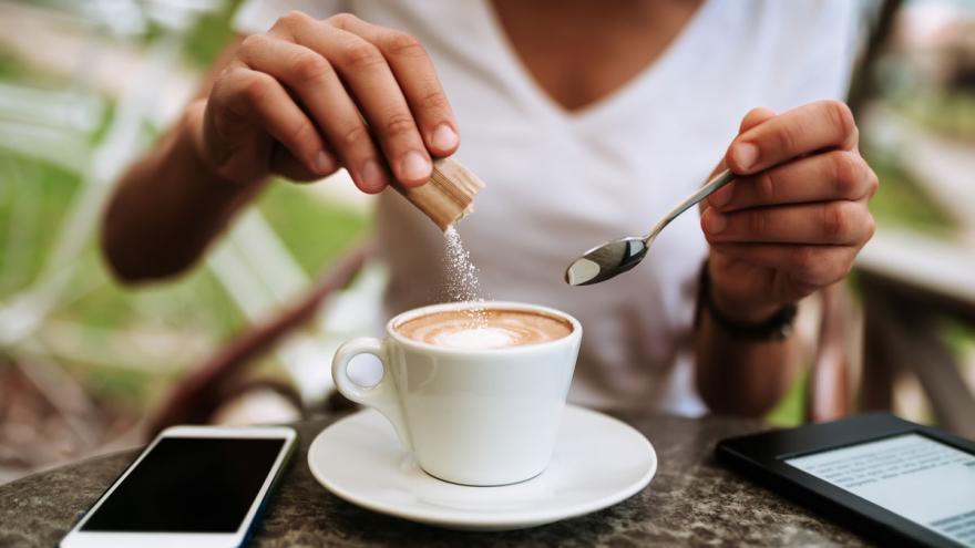 Junge Frau schüttet ZUcker in eine Tasse Cappuccino