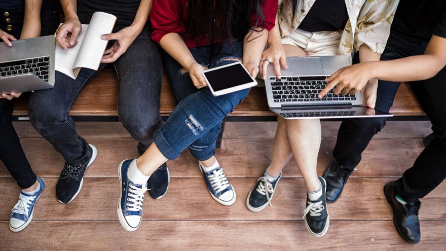 Junge Menschen sitzen auf einer Bank, Laptops und Tablets auf dem Schoß. Man sieht nur ihre Beine. 