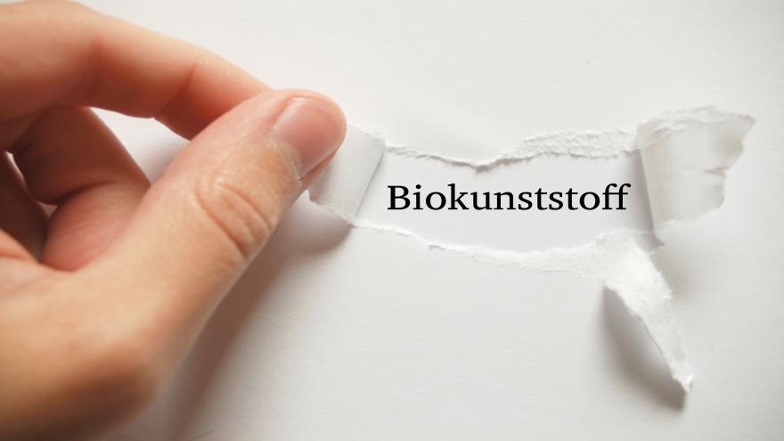 Finger, die ein Papier aufreißen, so dass das Wort Biokunststoff erscheint.