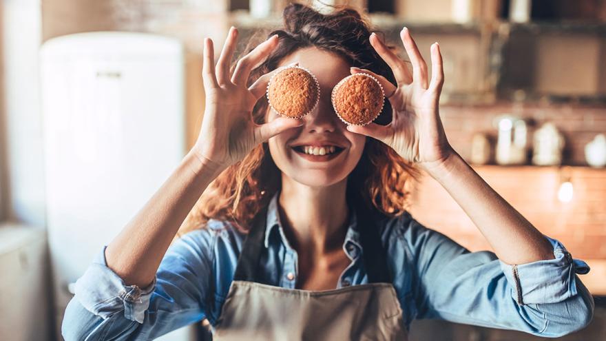 Eine junge Frau mit Kochschürze steht lachend in einer Küche und hält sich zwei Muffins vor die Augen 