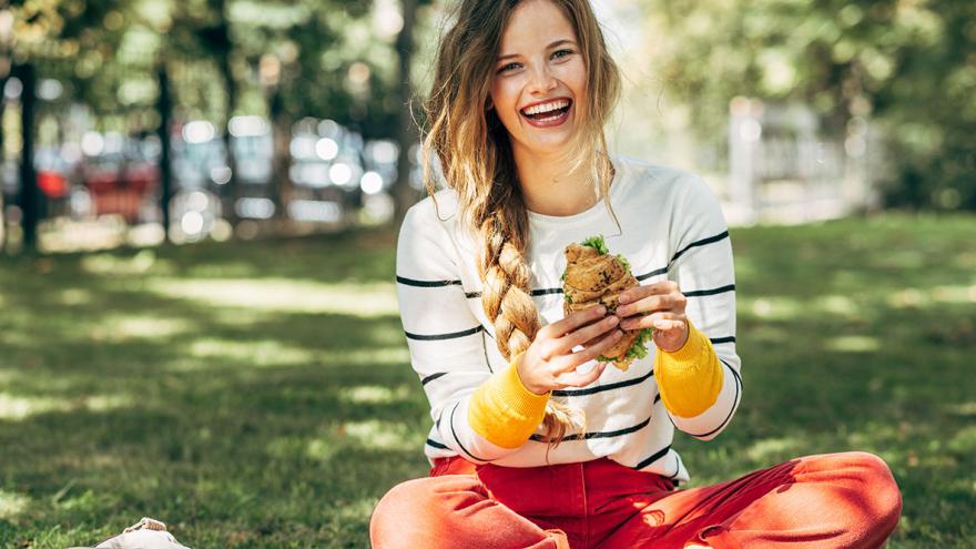Eine lachende junge Frau sitzt mit einem Brötchen in der Hand auf einer Wiese