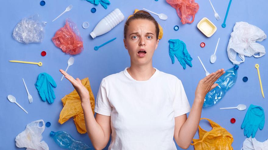 Eine junge Frau steht vor einer hellblauem Wand, die mit vielen Einweg-Plastikartikeln wie Besteck, Flaschen, Handschuhe, Tüten und Essensverpackungen bestückt ist 