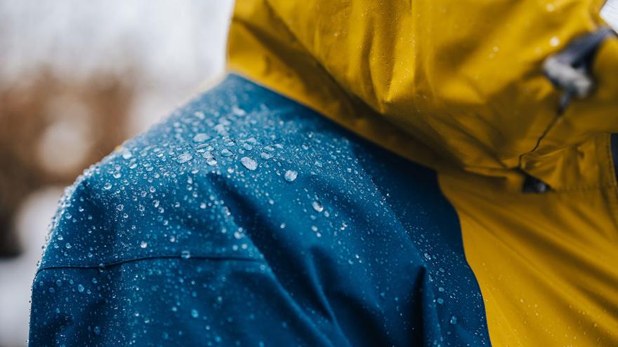 Jemand trägt eine blau-gelbe wasserabweisende Regenjacke mit abperlenden Regentropfen auf der Schulter