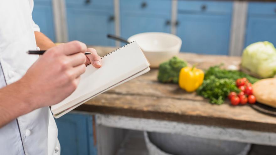 Jemand steht in einer Küche und macht sich Notizen, im Hintergrund eine Ablage mit Gemüse