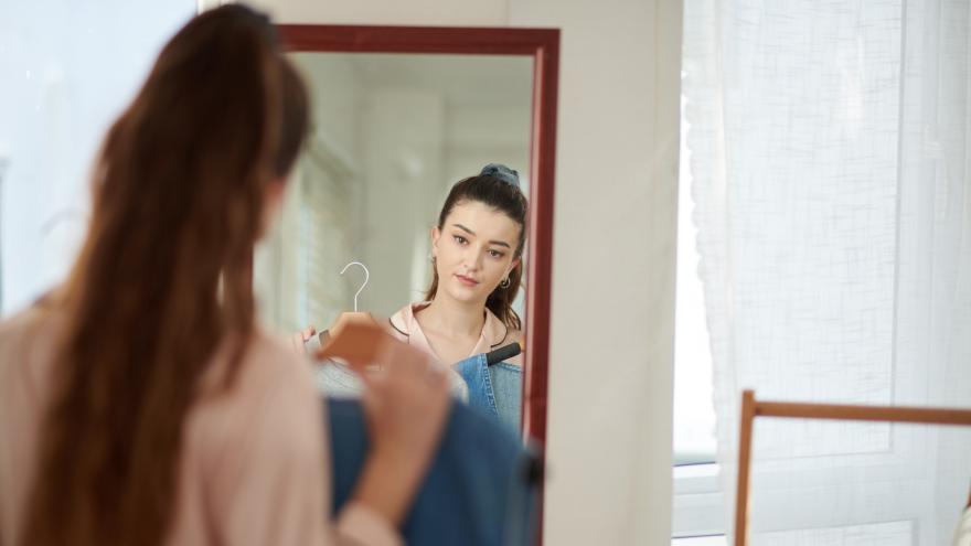 Eine junge Frau mit braunem Haar steht vor einem Spiegel und hält verschiedene Kleidungsstücke auf Bügeln vor ihren Körper. Ihr Gesicht ist in der Spiegelreflexion deutlich zu sehen, während sie prüfend in den Spiegel blickt.