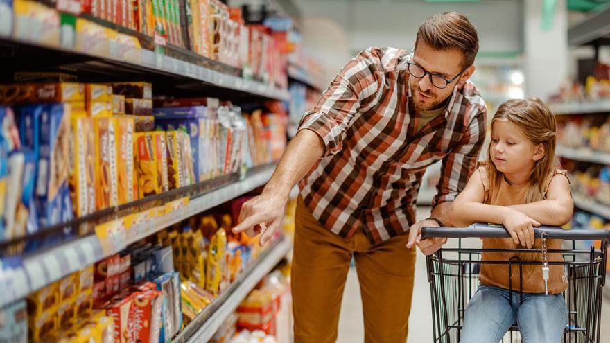 Ein Mann im Supermarkt deutet auf Produkte im Regal, eine Kind sitzt im Einkaufwagen daneben,  