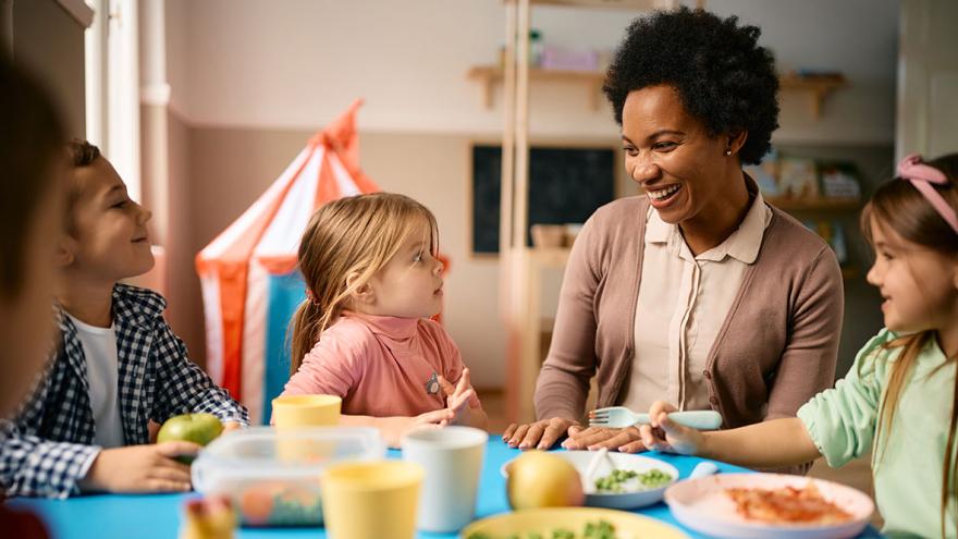 Eine Erzieherin sitzt mit Kindern im Kindergartenalter am Esstisch 