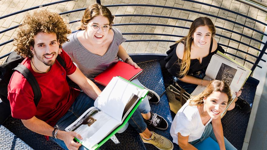 Vier junge Erwachsene sitzen mit Büchern auf einer Wendeltreppe und schauen nach oben in die Kamera 