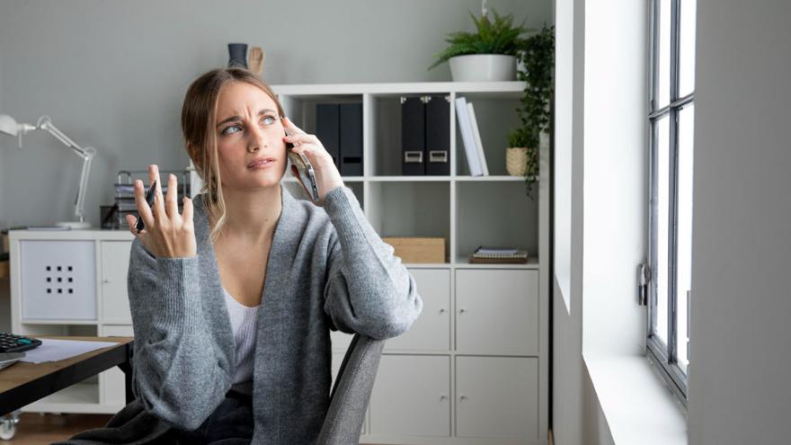Eine Frau sitzt genervt mit dem Telefon am Ohr an einem Schreibtisch 