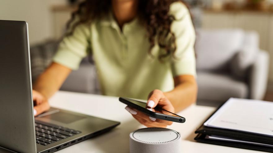 Eine Frau sitzt am Schreibtisch vor einem Laptop, ein Smartphone in der Hand