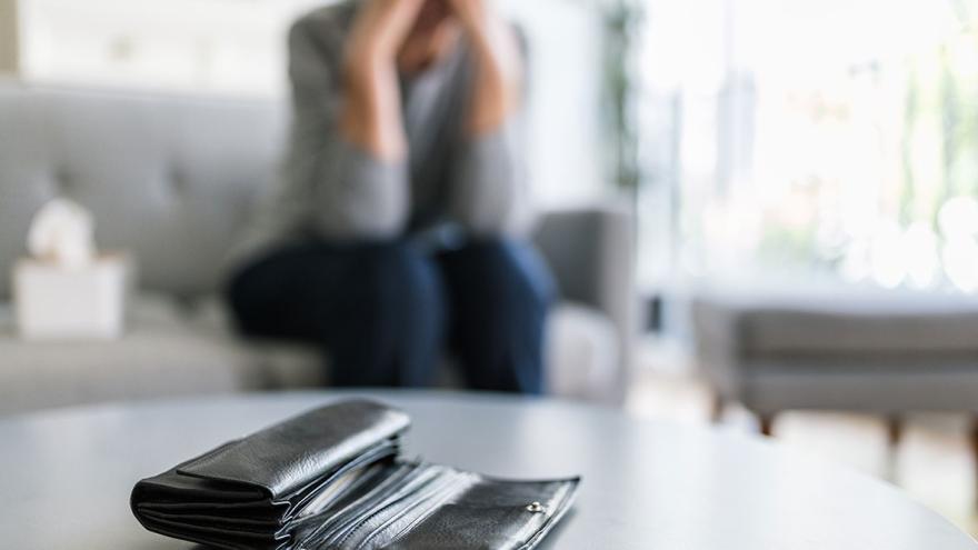 Jemand sitzt verzweifelt auf einem Sofa, im Vordergrund eine geöffnete Geldbörse auf einem dem Tisch