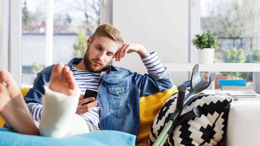 Ein junger Mann liegt verletzt mit eingegipstem Bein auf dem Sofa und schaut auf sein Smartphone