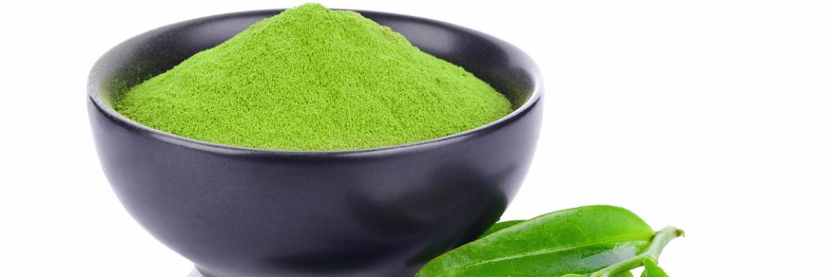 Grünes Matcha-Pulver in einer schwarzen Keramikschale und Teeblätter davor.