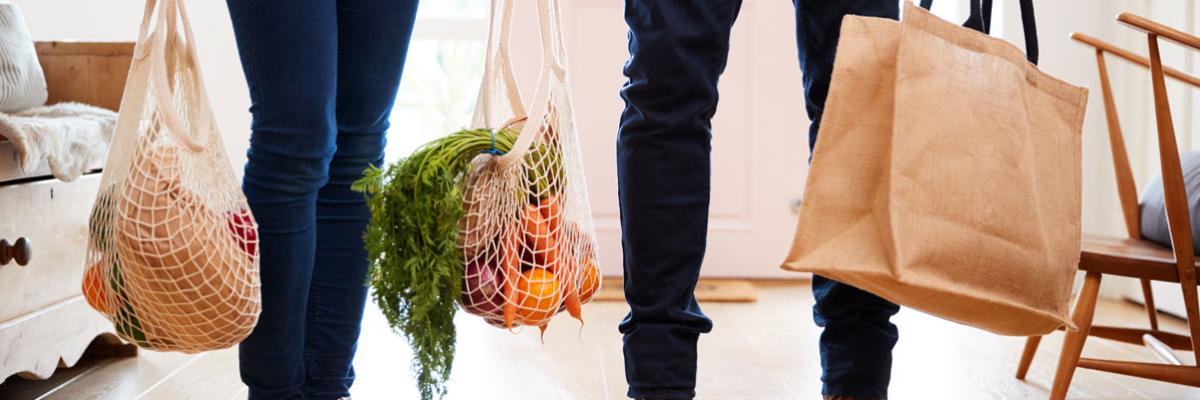 Zwei Personen mit Lebensmitteln in Einkaufsnetzen und -taschen stehen in einer Küche, man sieht nur ihre Beine