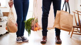 Zwei Personen mit Lebensmitteln in Einkaufsnetzen und -taschen stehen in einer Küche, man sieht nur ihre Beine
