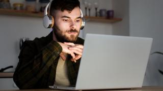 Ein junger Mann sitzt mit Headset vor einem Laptop am Küchentisch 