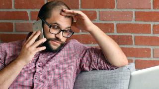 Ein Mann sitzt genervt mit seinem Smartphone am Ohr vor seinem Laptop 