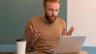 Ein Mann sitzt vor einem Laptop und hebt verärgert die Hände