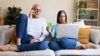 Ein Mann und eine Frau sitzen auf dem Sofa, der Mann telefoniert, die Frau hält erschrocken eine Kreditkarte hoch 
