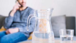 Im Hintergrund sitzt ein Mann telefonierend auf dem Sofa, vor ihm eine Karaffe und ein Glas mit Wasser 