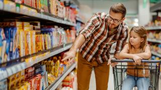 Ein Mann im Supermarkt deutet auf Produkte im Regal, eine Kind sitzt im Einkaufwagen daneben,  