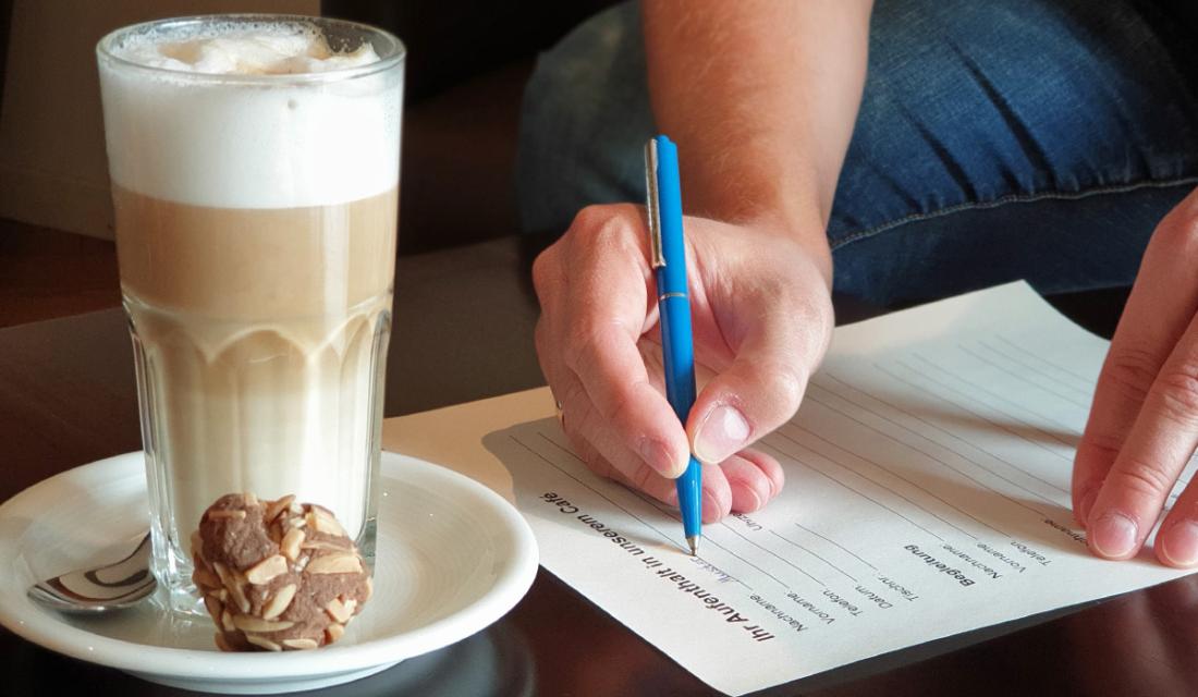 Kaffee und Gebäck auf einem Tisch, daneben schreibt jemand mit Kugelschreiber auf ein Blatt Papier